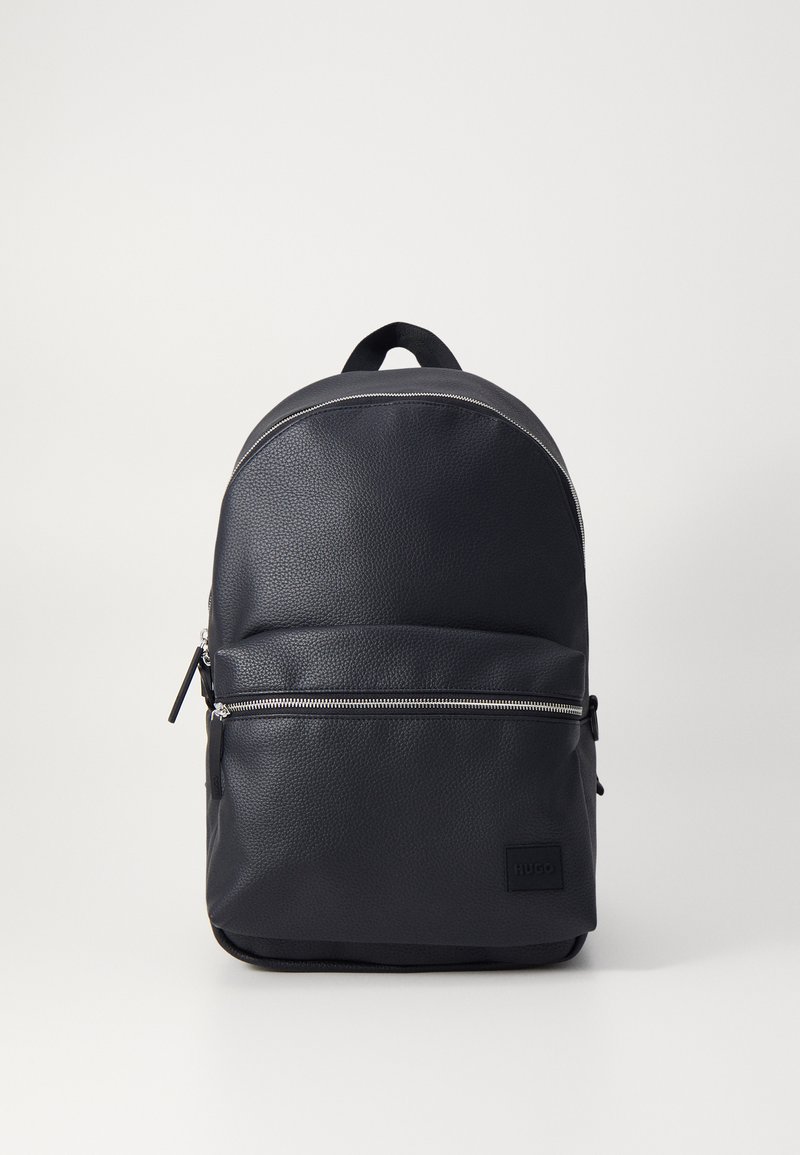 Mochila de cuero negro con un acabado suave, parte superior redondeada, bolsillo frontal con cremallera plateada y un parche de logo en la parte inferior.