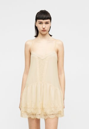 Jauna moteris su tiesiais tamsiais plaukais dėvi smėlio spalvos suknelę be rankovių, puoštą nėriniais, stovinti priešais vienodu baltu fonu.