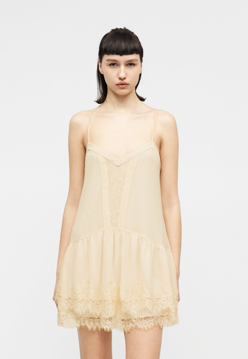 Νεαρή γυναίκα με ίσια σκούρα μαλλιά, φορώντας μπεζ αμάνικο φόρεμα με δαντελωτό τελείωμα, μπροστά σε απλό λευκό φόντο.