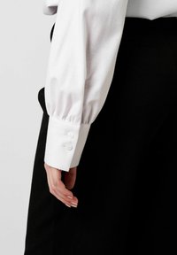 Weiße Langarmbluse mit Puffärmel, die zwei Knopfdetails aufweist, kombiniert mit schwarzem Stoff. Nahaufnahme des Ärmels und der Hand.