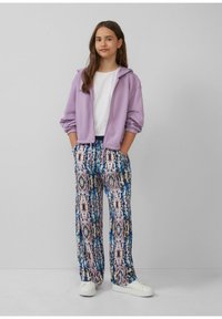 Lilafarbene Zip-Up-Hoodie über einem weißen T-Shirt, kombiniert mit weit geschnittenen, bunten gemusterten Hosen in Blau, Pink und Braun. Weiße Sneaker.