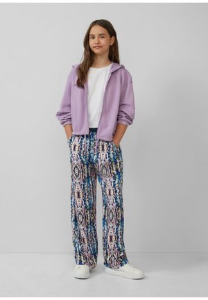 Un sweat à capuche lilas zippé sur un t-shirt blanc, associé à un pantalon large à motifs colorés avec des accents bleus, roses et bruns. Des baskets blanches.