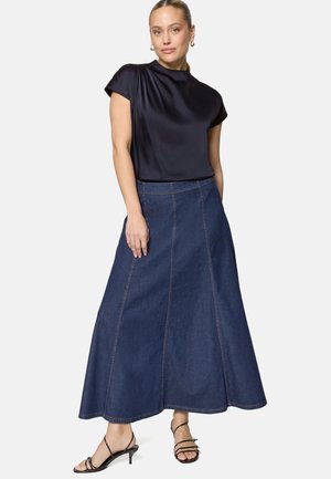 Kobieta ubrana w czarną satynową bluzkę z krótkim rękawem, długą ciemną jeansową spódnicę w kształcie litery A oraz czarne sandały na obcasie z paskami na tle jednolitego tła.