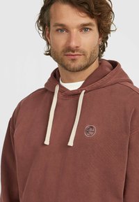 Bruine hoodie van zacht materiaal met crèmekleurige trekkoorden, voorzien van een rond logo op de borst en een relaxte pasvorm.