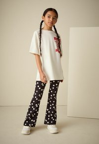 T-shirt blanc oversize avec une impression graphique rouge, associé à un pantalon noir évasé orné de gros pois blancs. Des baskets blanches complètent la tenue.