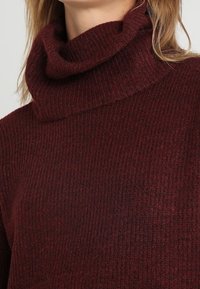 Maglione a collo alto color marrone scuro realizzato in tessuto a maglia testurizzato. Presenta un colletto piegato e un motivo a coste, che enfatizzano calore e comfort.