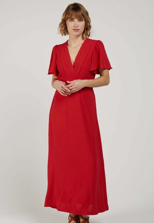Femme aux cheveux courts et ondulés portant une robe maxi rouge à col en V avec des manches courtes volantées, debout les mains jointes.