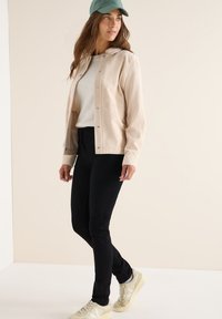 Beige kordjacka med tryckknappar, bärs över en krämfärgad tröja och svarta jeans. Avslutad med en grön keps och ljusa sneakers.