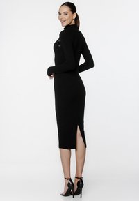 Czarna, prążkowana sukienka dzianinowa z długimi rękawami, wysokim kołnierzem, bocznym rozcięciem oraz guzikami z tyłu. Noszona z czarnymi sandałami na wysokim obcasie.