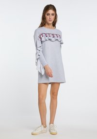Graues Sweatshirt-Kleid mit einem gerafften Frontdetail, das lila Akzente aufweist. Lange Ärmel und eine lässige Passform, kombiniert mit weißen Sneakers.