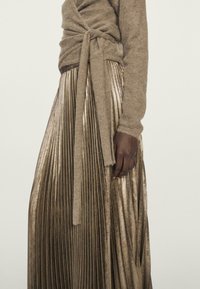 Beiger Strickpullover mit Kordelzug in der Taille; plissierter metallic goldener Rock mit glänzender Textur. Das Outfit zeichnet sich durch kontrastierende Materialien aus.