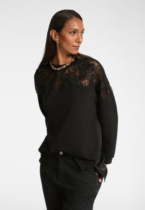 Vrouw in een zwart top met lange mouwen en een ingewikkelde kant yoke, met gestreepte zwarte broek, gouden ketting en gouden creolen, kijkt naar rechts.