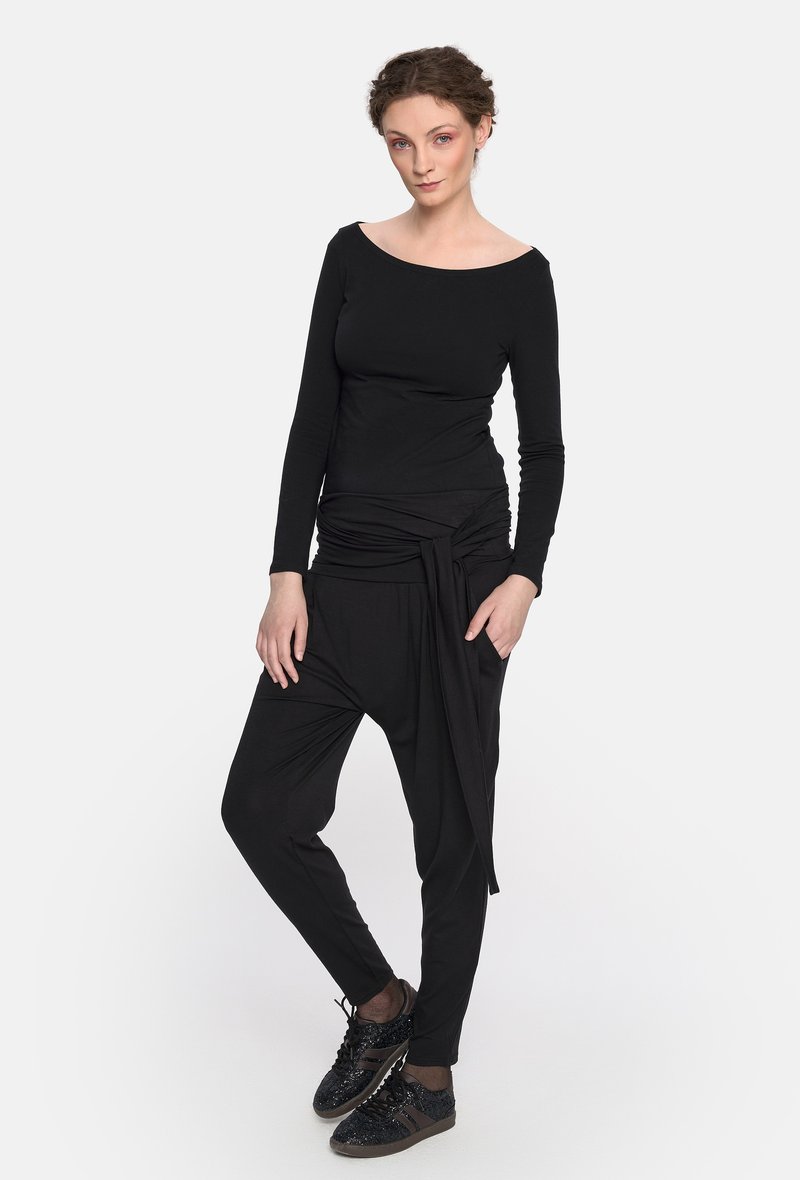 Czarny top z długim rękawem o dekolcie odkrywającym ramiona, w połączeniu z luźnymi czarnymi spodniami z wiązanym detalem w talii i czarnymi sneakerami.