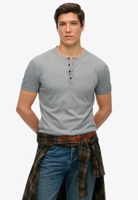 Szara koszulka henley z krótkim rękawem i trzema czarnymi guzikami, w połączeniu z niebieskimi dżinsami oraz zieloną i brązową koszulą w kratę owiniętą wokół talii.