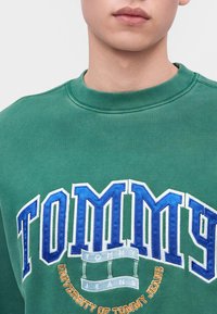Човек, носещ зелен суитшърт с големи сини и бели "TOMMY" букви и по-малък текст "University of Tommy Jeans", бродиран по-долу.