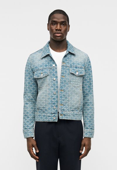 Άνδρας που φοράει ανοιχτό μπλε τζιν μπουφάν με σχέδιο πάνω από λευκό πουκάμισο και μαύρο παντελόνι, στέκεται μπροστά σε απλό φωτεινό φόντο.