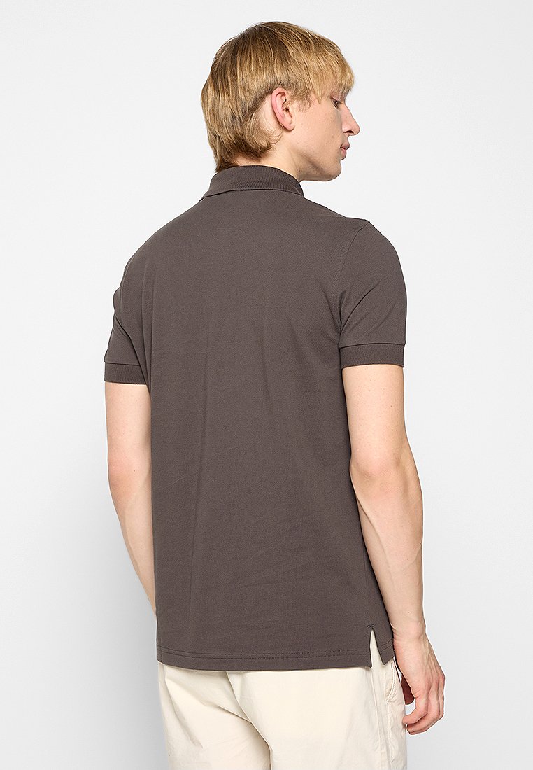 Homme portant un polo à manches courtes marron foncé et un pantalon beige clair, se tenant de dos avec la tête tournée sur le côté.