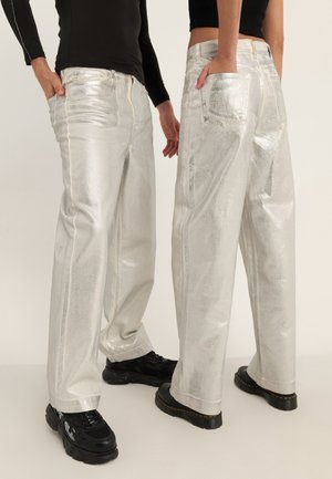 Dwie osoby w błyszczących srebrnych szerokich spodniach i czarnych topach, stojące z rękami w kieszeniach, widoczne czarne masywne buty.