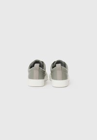 Sapatos desportivos verdes com um acabamento liso e texturizado, solas brancas contrastantes e detalhes em tecido cinza claro na parte de trás.