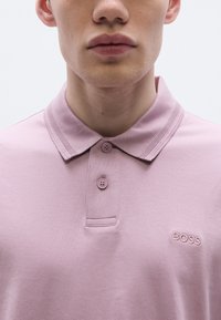 Roza polo majica s klasičnim ovratnikom, z dvema gumboma na sprednji strani in izvezenim logotipom BOSS v ujemajoči se barvi na levi strani prsnega koša.