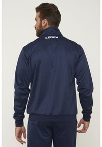 Chaqueta deportiva azul marino de tejido suave, con cuello alto y puños acanalados. En la parte trasera presenta el logo "LEGEA" en blanco.