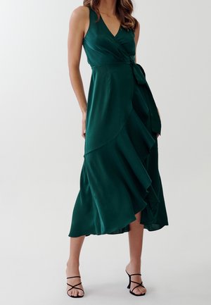 Vestido tipo wrap en satén verde oscuro, con un escote en V profundo, cinturilla ajustable con lazo, dobladillo asimétrico con volantes y elegantes tacones finos negros.