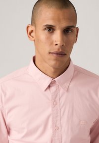 Camisa de botones rosa claro con un cuello, que presenta un логотип sutil en el lado izquierdo del pecho y una tela suave y texturizada.