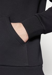 Sudadera negra con un bolsillo frontal y puños acanalados, que presenta una textura suave y un diseño ajustado. Sin patrones ni logotipos visibles.