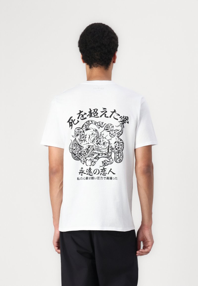 Hvid bomulds t-shirt med et sort grafisk design på ryggen, der viser intrikate kunstværker og japansk tekst. Korte ærmer og rund hals.