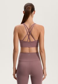 Vijolični športni modrček z križanjem trakov na zadnji strani. Gladka tekstura in podporna zasnova. OYSHO logo na spodnjem traku.