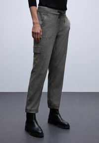 Pantalons de jogging gris en tissu texturé, dotés d'une taille élastique avec cordon de serrage et de poches cargo latérales. Des bottines noires sont portées.