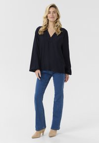 Blusa a maniche lunghe blu navy con scollatura a V e vestibilità ampia, abbinata a jeans aderenti blu chiaro e scarpe con tacco beige. Colore uniforme, dettagli minimi.