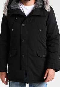 Parka noire avec capuche bordée de fourrure, fermeture par boutons et zip, deux poches avant et doublure intérieure douce et texturée.