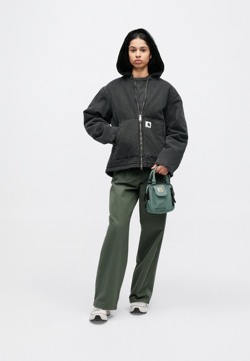 Schwarze Kapuzenjacke mit einem Frontreißverschluss und Seitentaschen, kombiniert mit grüner Hose und einer kleinen türkisfarbenen Umhängetasche. Weiße Sneakers runden den Look ab.