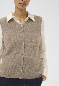 Beige strikket cardigan med V-udskæring, med knaplukning og ribbet tekstur, lagdelt over en cremefarvet skjorte med blomster mønster.