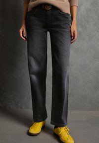 Persona con jeans rectos de color gris oscuro, un suéter marrón y zapatillas brillantes amarillas, de pie sobre un suelo gris.