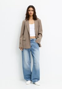 Blazer beige ajusté, débardeur blanc court et jeans bleu clair amples avec des détails déchirés. Le mannequin porte des baskets blanches.