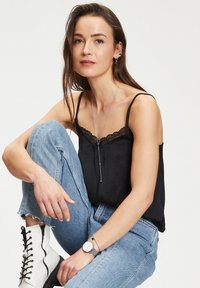 Sort spaghetti top med spidsede stropper og blonde detaljer, parret med lyseblå slidte jeans og hvide snøre støvler. Accessoriseret med et ur.