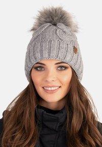 Grijze gebreide beanie met een gestructureerd patroon, afgewerkt met een fluffy grijze pompon. Heeft een klein leren accentlabel aan de zijkant.