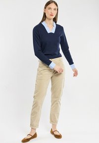 Marineblauwe v-halstrui met lichtblauwe kraag en manchet, gecombineerd met beige corduroy broeken en bruine platte schoenen.