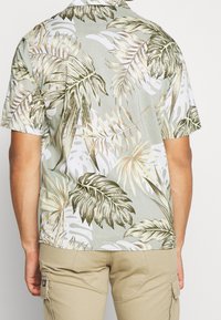Mies, joka on pukeutunut vaaleisiin cargo-housuihin ja lyhythihaiseseen paitaan, jossa on vaaleanvihreitä, valkoisia ja ruskeita trooppisia lehtikuvioita, nähtynä takaa.