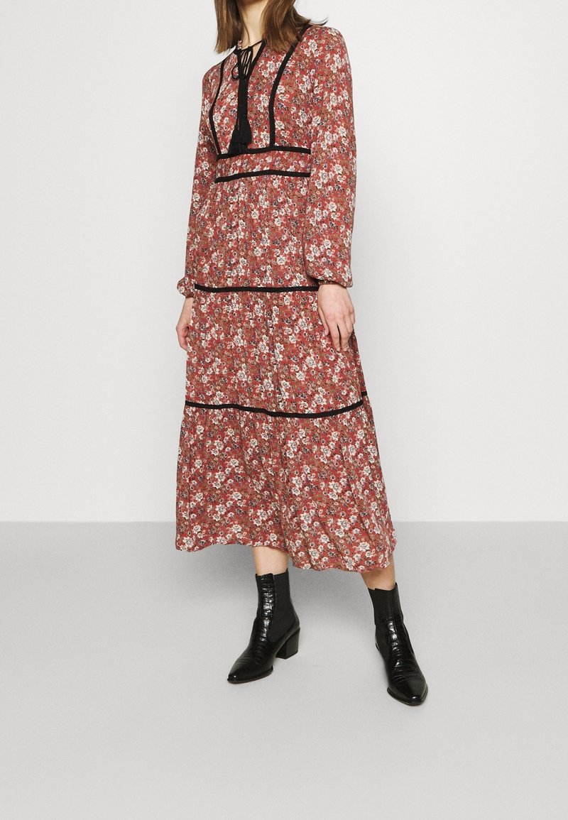 Rotes Maxikleid mit langen Ärmeln, das mit schwarzem Besatz und einem gebundenen Hals ausgestattet ist. Das Material ist leicht und bietet eine fließende Silhouette sowie ein gestuftes Design.