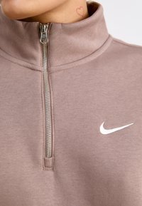Felpa zip-up marrone chiaro con colletto alto, caratterizzata da un logo Nike bianco e un tessuto testurizzato. Dettaglio della cerniera in argento al collo.