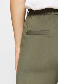 Olivgrüne Hose mit einem elastischen Bund und einer einzigen Gesäßtasche mit einer genähten Akzentuierung. Glatte, matte Textur.