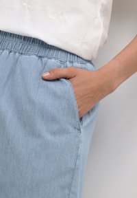 Vaaleansiniset farkut, joissa on joustava vyötärönauha, käsi taskussa, joka näyttää sileän rakenteen ja rentoa istuvuutta. Valkoinen yläosa, jossa on hillitty kirjonta.