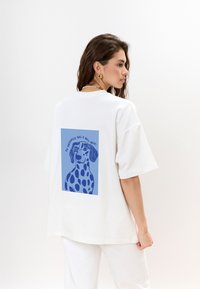 Witte oversized t-shirt met een blauwe grafische illustratie van een hond met vlekken op de rug, aangevuld met tekst in een gebogen formaat.