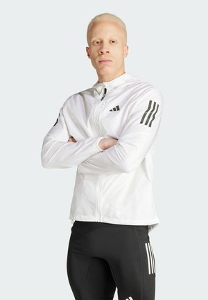Chaqueta deportiva blanca con cremallera en la parte delantera, diseñada con tres franjas negras en las mangas. Llevada sobre pantalones ajustados negros, que muestran un ajuste estilizado.