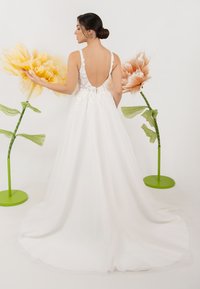 Weißes Hochzeitskleid mit Spitzenverzierungen, V-Ausschnitt und fließendem Rock. Blumenarrangements in lebhaften Gelb- und Pfirsichtönen im Hintergrund.