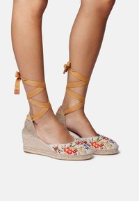 Espadrilky na klinovom podpätku s prírodnou juteovou podrážkou a vyšívaným kvetinovým dizajnom v červenej a fialovej farbe. Hnedé šnúrky sa omotávajú okolo členku.