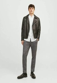 Jovem em pé vestindo uma jaqueta de couro preta, camisa branca, calças cinza xadrez, meias pretas e mocassins pretos contra um fundo simples.
